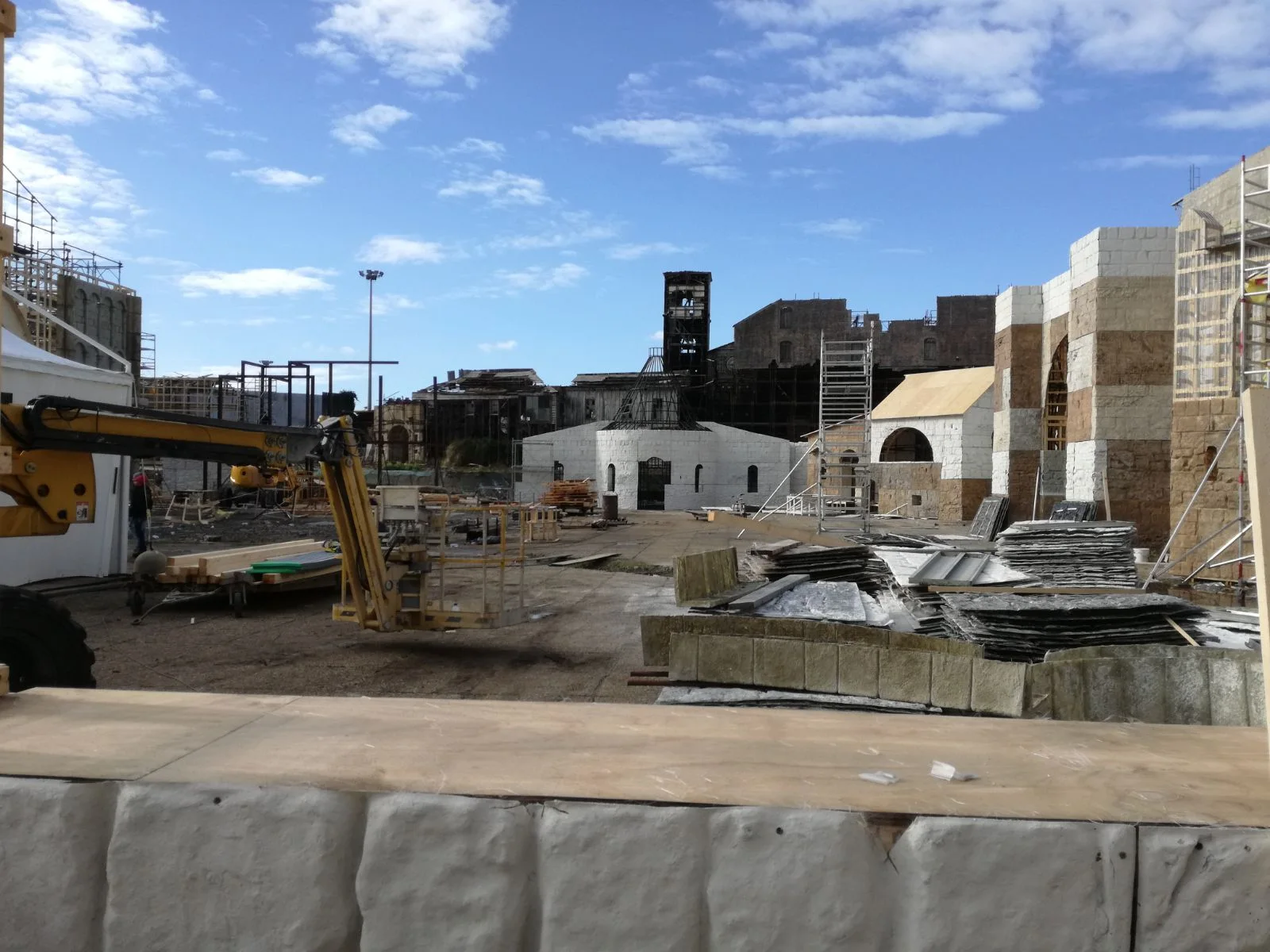 Vista grandangolare del backlot esterno: cantiere su terreno aperto con campanile dell'abbazia ancora a metà costruzione, gru, edifici monastici, ponteggi. Cielo azzurro con nuvole bianche