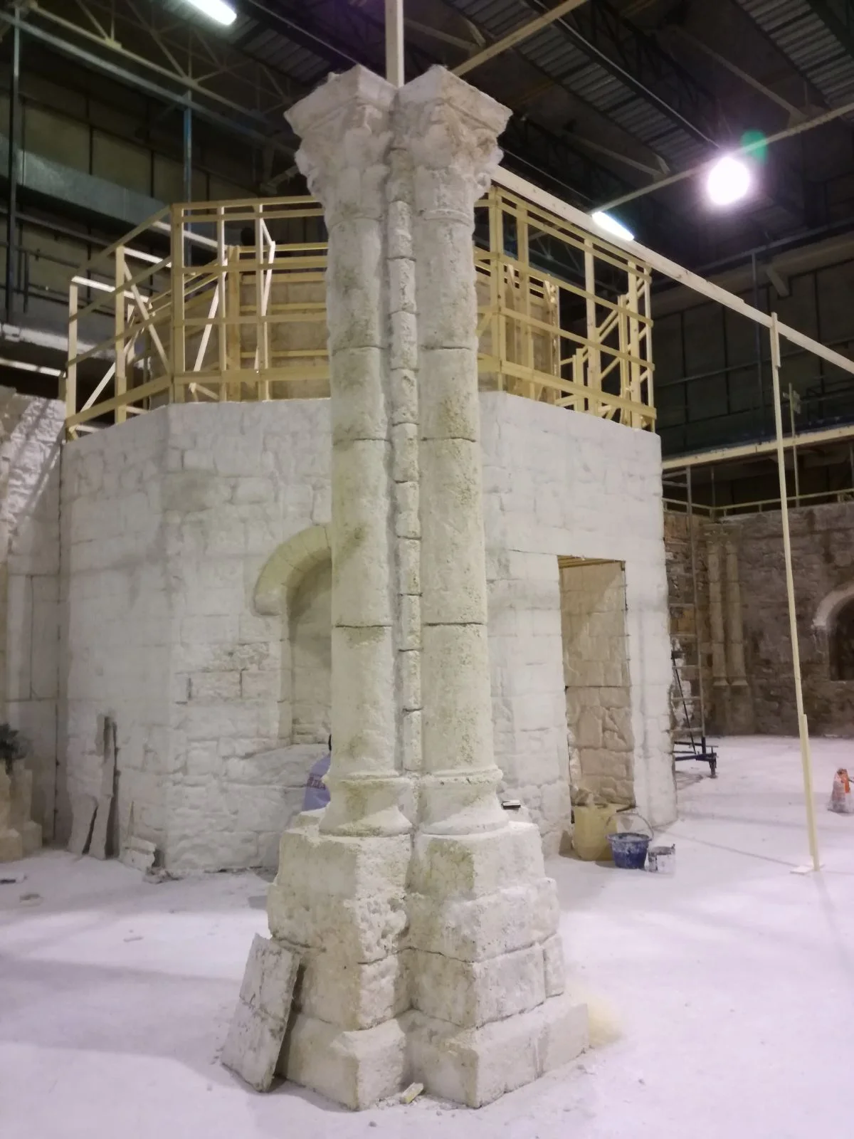 Una colonna gemellata in pietra finta scolpita a tutta altezza nel teatro di posa, capitelli a foglie d'acanto, base a doppio bulbo, ponteggi sullo sfondo