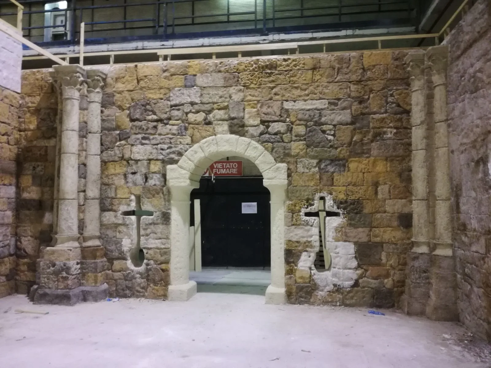 Portale ad arco a tutto sesto interno con muratura in pietra a vista e croci scolpite simmetriche sui due lati. Cartello rosso 'VIETATO FUMARE' affisso sopra la porta