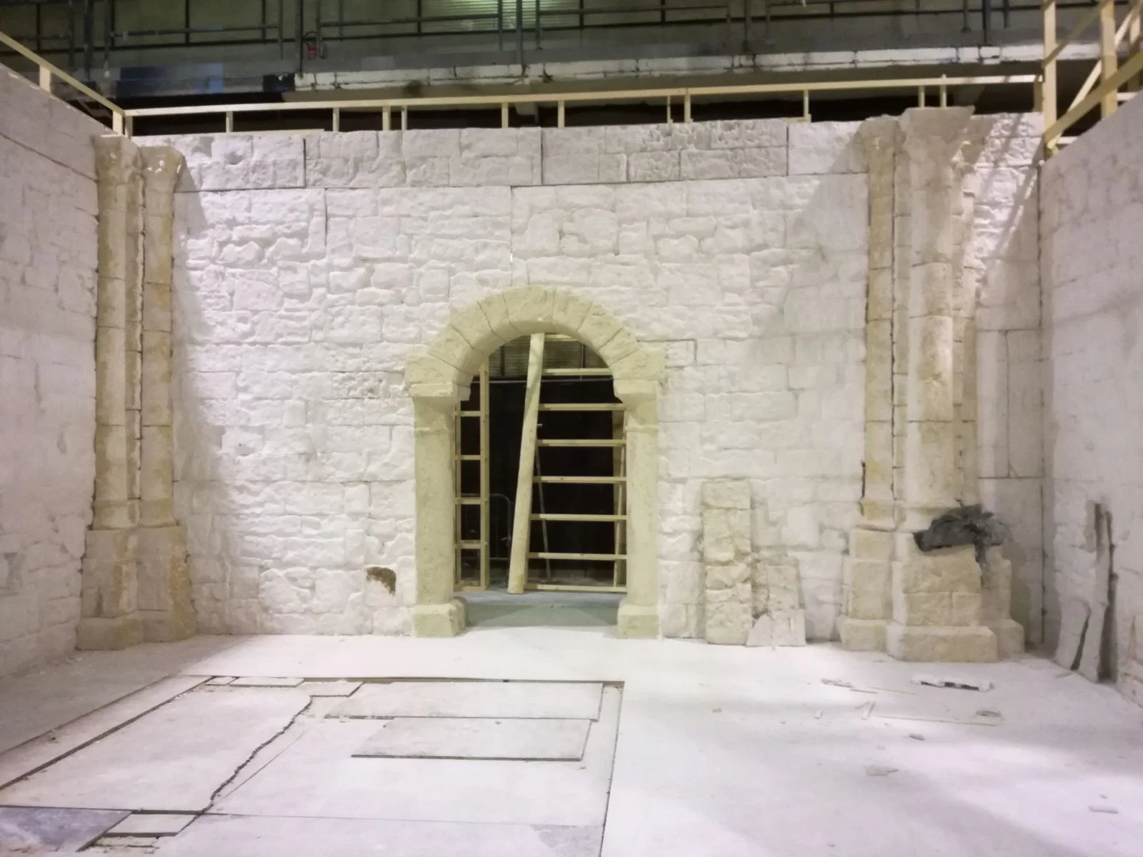 Un portale ad arco a tutto sesto in finta pietra, completato in studio: cornice gialla in pietra finta, parete di pietra a vista, ponteggio a destra. Set di interno abbazia in costruzione.