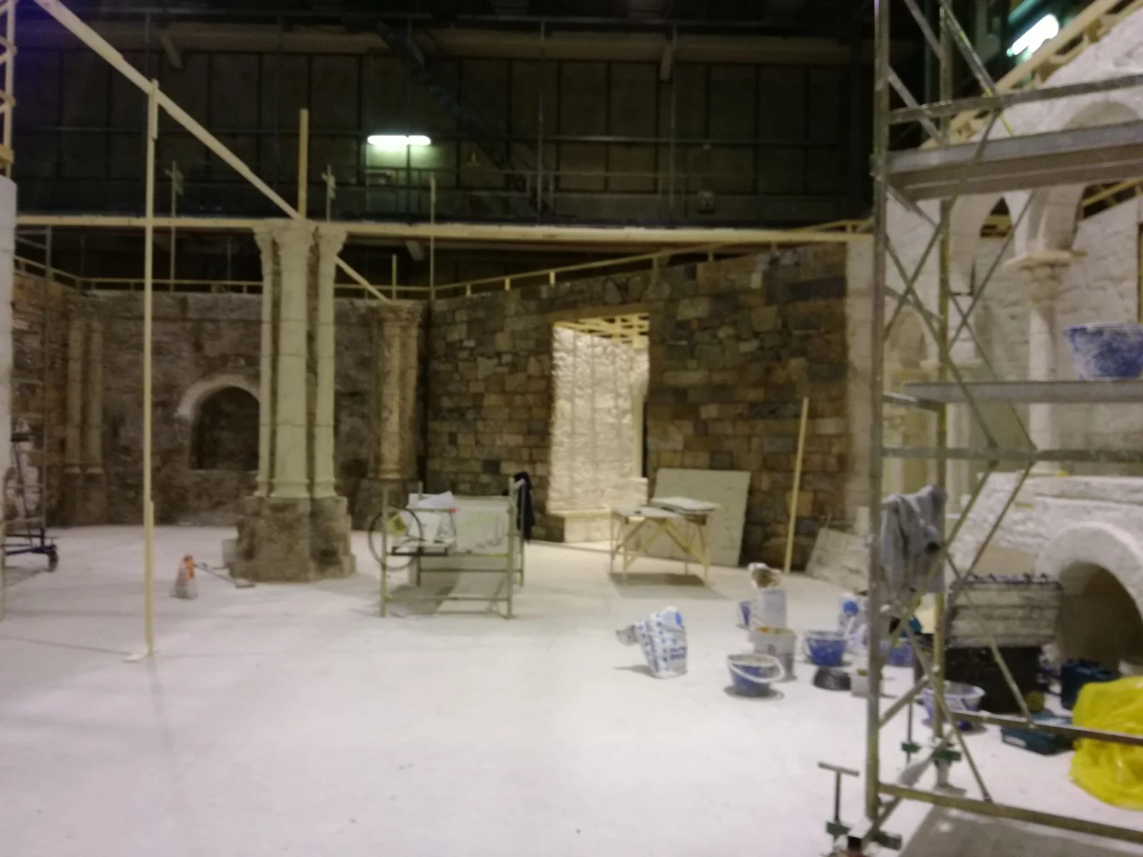 Vista d'insieme del set interno dell'abbazia in costruzione: muri in pietra mista, colonne gemellate, archi a tutto sesto, ponteggi a destra, secchi di malta blu in primo piano