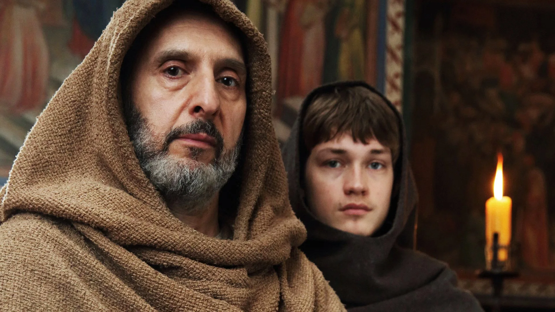 Primo piano di Guglielmo (John Turturro) con il cappuccio del saio francescano alzato e Adso (Damian Hardung) alle sue spalle. Sullo sfondo affreschi medievali a colori caldi.