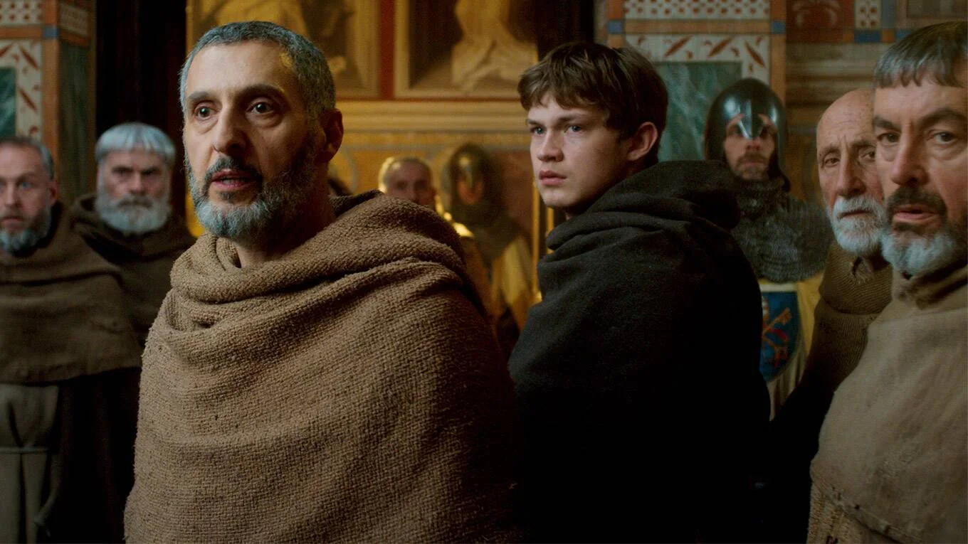 John Turturro nei panni di Guglielmo da Baskerville e Damian Hardung nei panni di Adso, circondati da monaci e da cavalieri armati con cotte di maglia. La sala ha pareti coperte di pitture parietali medievali, arazzi rossi
