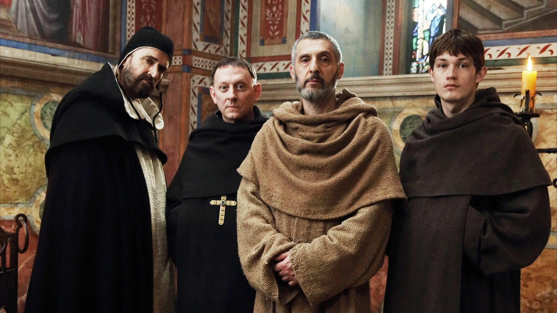 Quattro personaggi monastici della serie — Bernardo Gui, Severino, Guglielmo, Adso — in fila contro la parete affrescata della cappella, vetrata policroma a destra, candelabro acceso