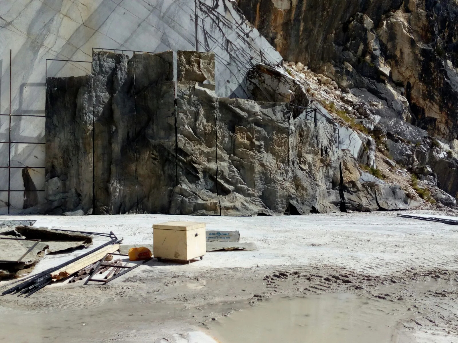 Worksite at the foot of the Carrara quarry: set construction in progress on the grey-white wall, foreground a white wooden crate for equipment, marble fragments and metal scaffolding on the left, ground white with marble dust
