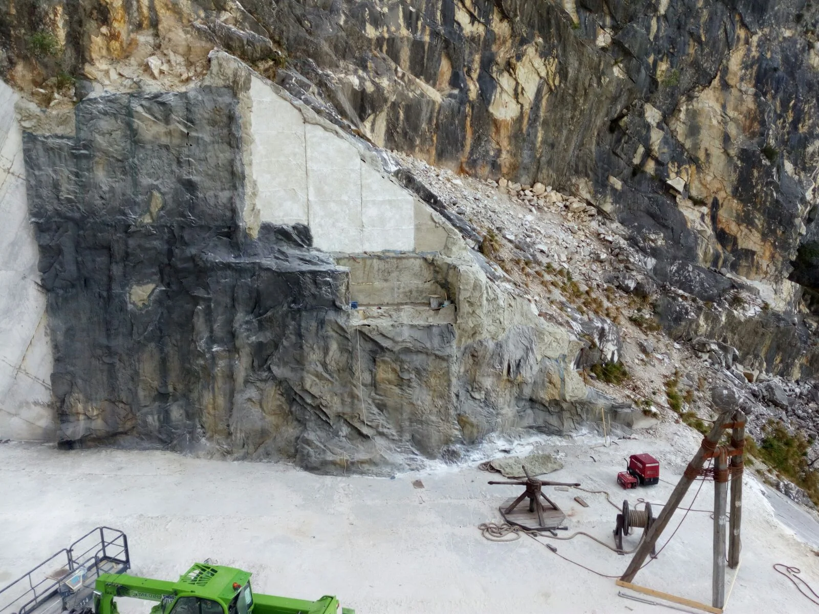 Panoramic view of the worksite inside the Carrara quarry: the great white-grey wall of the active quarry, at the centre the scenographic intervention with triangular panels, lower left a green excavator, on the right a red industrial winch and a black drum winch used to lift the blocks