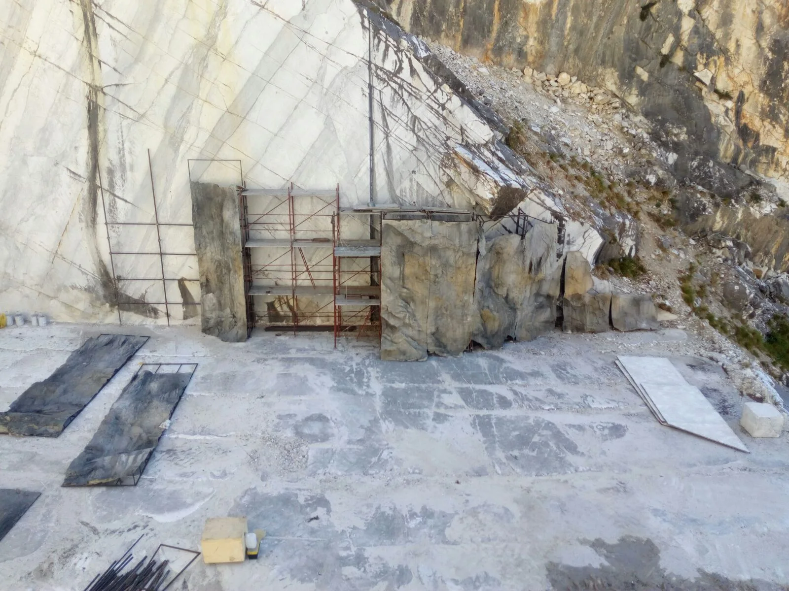 High-angle view of the worksite: metal scaffold hooked to the white quarry wall with applied panels, in the middle large real marble blocks resting on the yard, below black tarpaulins laid out for protection, on the right a narrow metal stair