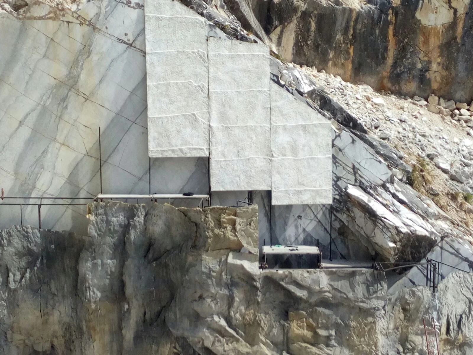 Close-up detail of the faux-marble panels in place: three overlapping white-grey triangular cusps stacked like steps, anchored to the real quarry wall with tie-rods, below a metal scaffold and a work platform