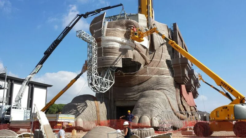 The elephant being worked seen front-on from low angle: enormous head, eye being finished, telescopic cranes on the sides with workers on the cherry-pickers