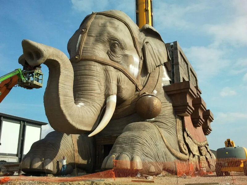 Finished elephant seen front-on with red ceremonial harness, golden bell, ivory-effect tusks, painted eyes, crane on the left still working on details