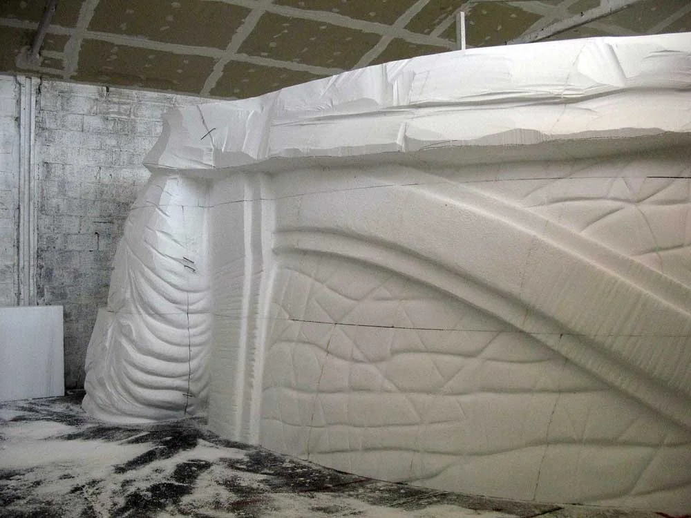 Piece of the Indiana Adventure elephant sculpted in raw white polystyrene in the Creation Art workshops: leg with detailed skin folds, front view, industrial warehouse in the background with white dust on the floor