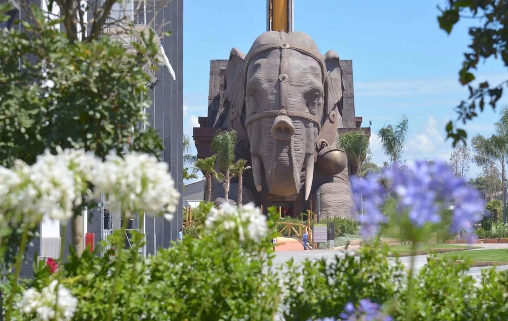 The Indiana elephant seen in daylight at the park, with palm trees around, the temple structure behind, visitors at the entrance