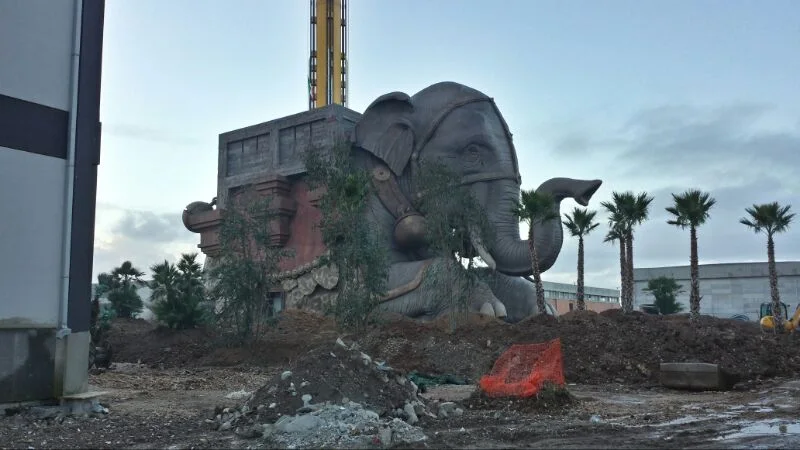 The elephant seen from medium distance under construction, palm trees already planted around, side red temple structure with oriental details