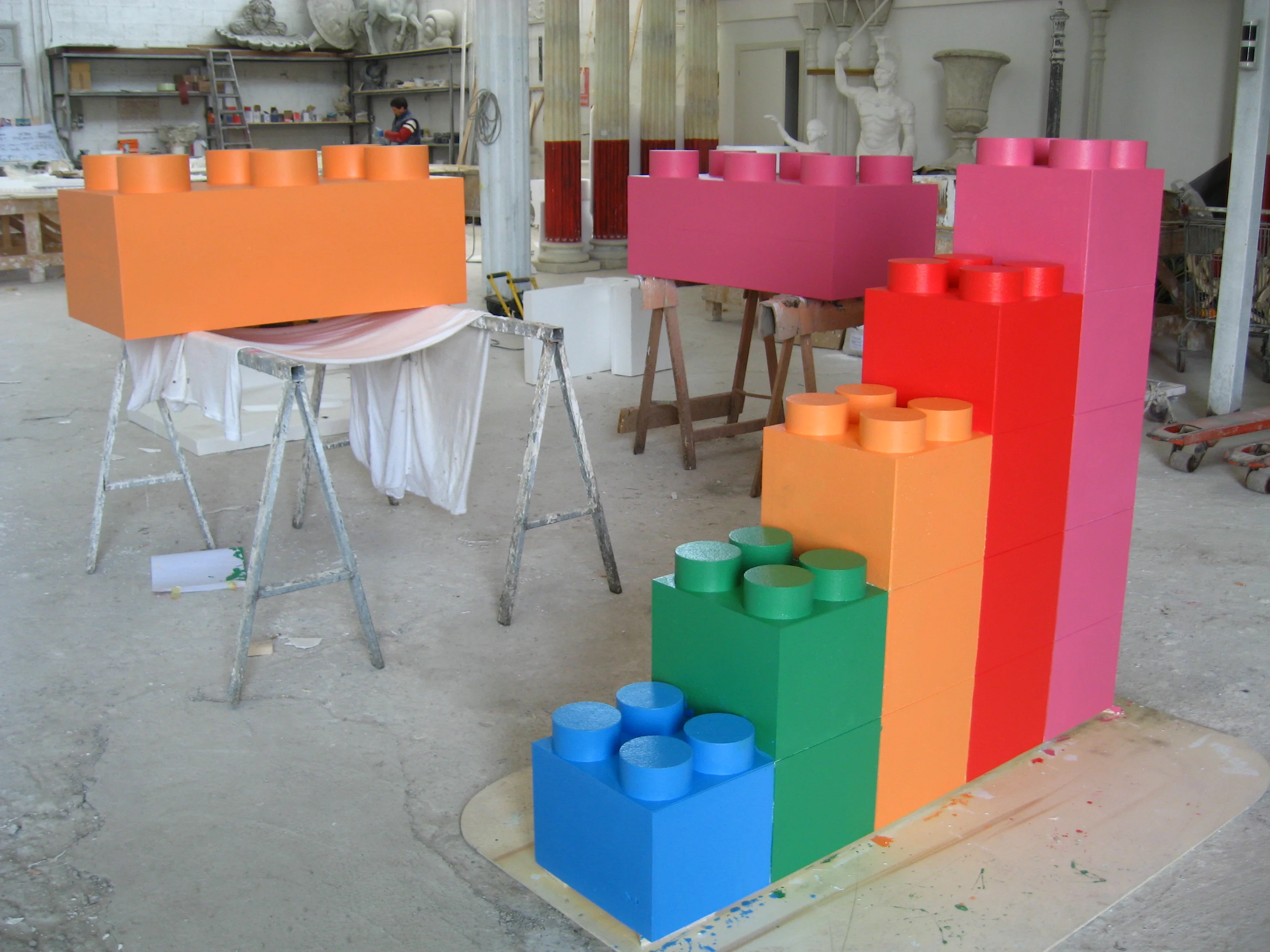 Set of giant scenographic LEGO bricks sculpted in our workshops for Italia's Got Talent: on the left a large orange LEGO brick laid horizontally on a painter's easel, on the right an ascending-staircase pyramid of polychrome bricks — a blue brick at the bottom, then green, then orange, then red, with pink and magenta on top; in the background the archive of classical statues in our workshop