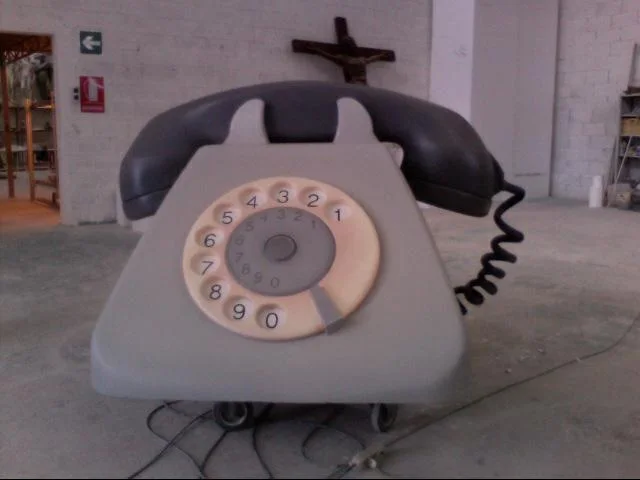 The same SIP Bigrigio phone finished in polychrome: light-grey base, anthracite-black handset, white dial with numbers from 0 to 9, black spiral cord trailing onto the workshop floor, a small crucifix on the wall above the phone