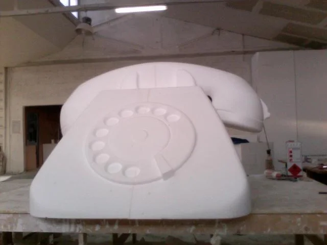 SIP Bigrigio landline phone sculpted in raw white polystyrene in our workshop: classic 1970s Italian phone shape with a square base and handset on top, stylised central rotary dial with concentric grooves, set on a workbench with the workshop walls in the background