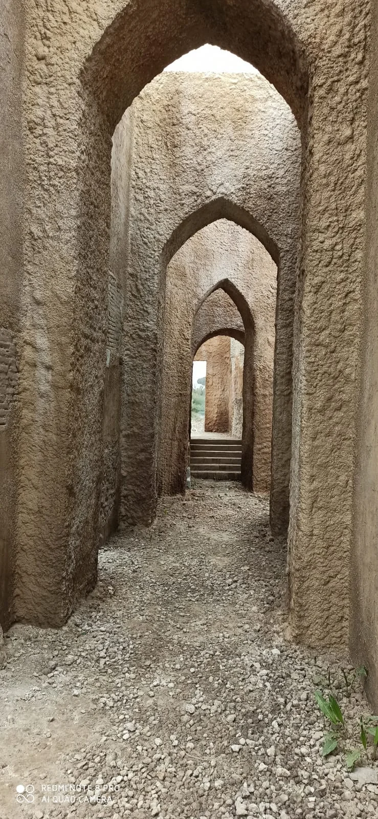 Corridoio-galleria con arcate gotiche a sesto acuto in finta pietra: sequenza di archi che si rimpiccioliscono verso il fondo come prospettiva forzata, pareti color pietra erosa con texture rugosa, ghiaione di terra battuta sul pavimento, in fondo una soglia con gradini di pietra che porta verso la luce