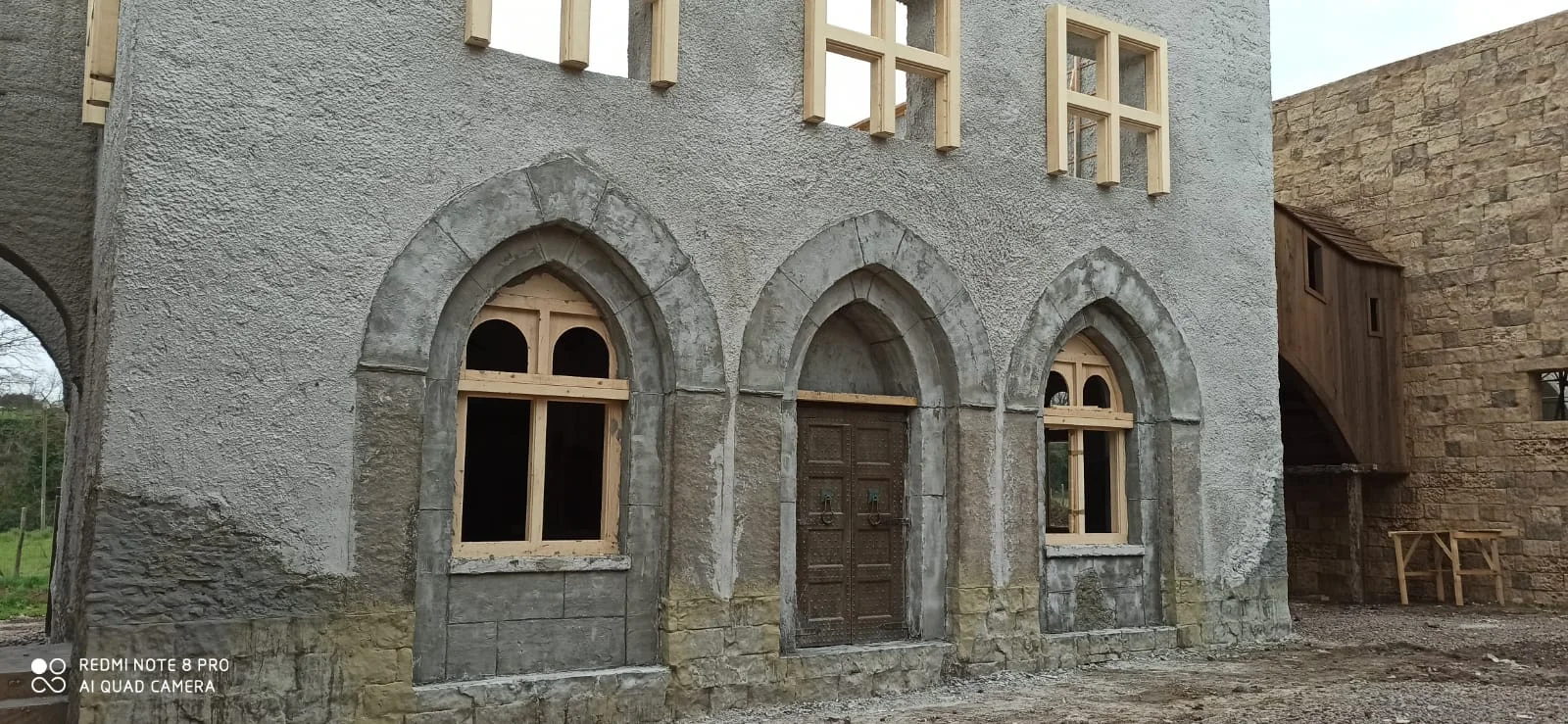 Facciata del palazzo lombardo a due piani: parete in finta pietra grigia bugnata al piano terra, tre archi gotici a sesto acuto con cornici in pietra chiara, porta centrale in legno scuro, finestre bifore di legno chiaro al piano superiore, parete superiore in intonaco grigio chiaro; alla destra un edificio attiguo in pietra di taglio chiaro