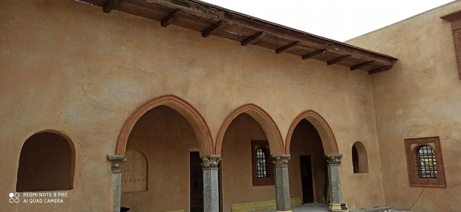 Portico a tre arcate ribassate: cornici degli archi in cotto rosso, colonne in pietra grigia con capitelli a foglie d'acanto stilizzati, parete in intonaco color sabbia-ocra modulato, sotto le arcate si vedono ingressi a porta scura e finestre con cornici cotto, sopra il portico tetto in travi di legno a vista e un'altra finestra ad arco