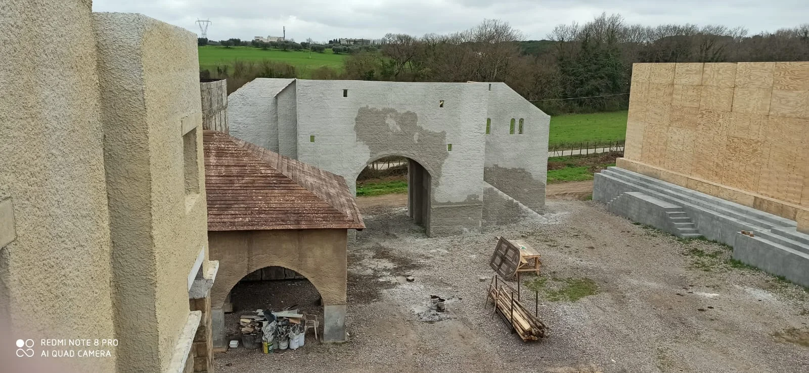 Vista panoramica del villaggio rinascimentale del set di Leonardo: due palazzi in finta pietra grigio-bianca con arco di passaggio centrale e tetto a tegole rosse, edifici secondari in legno chiaro sulla destra, paesaggio collinare verde sullo sfondo, cielo grigio nuvoloso primaverile