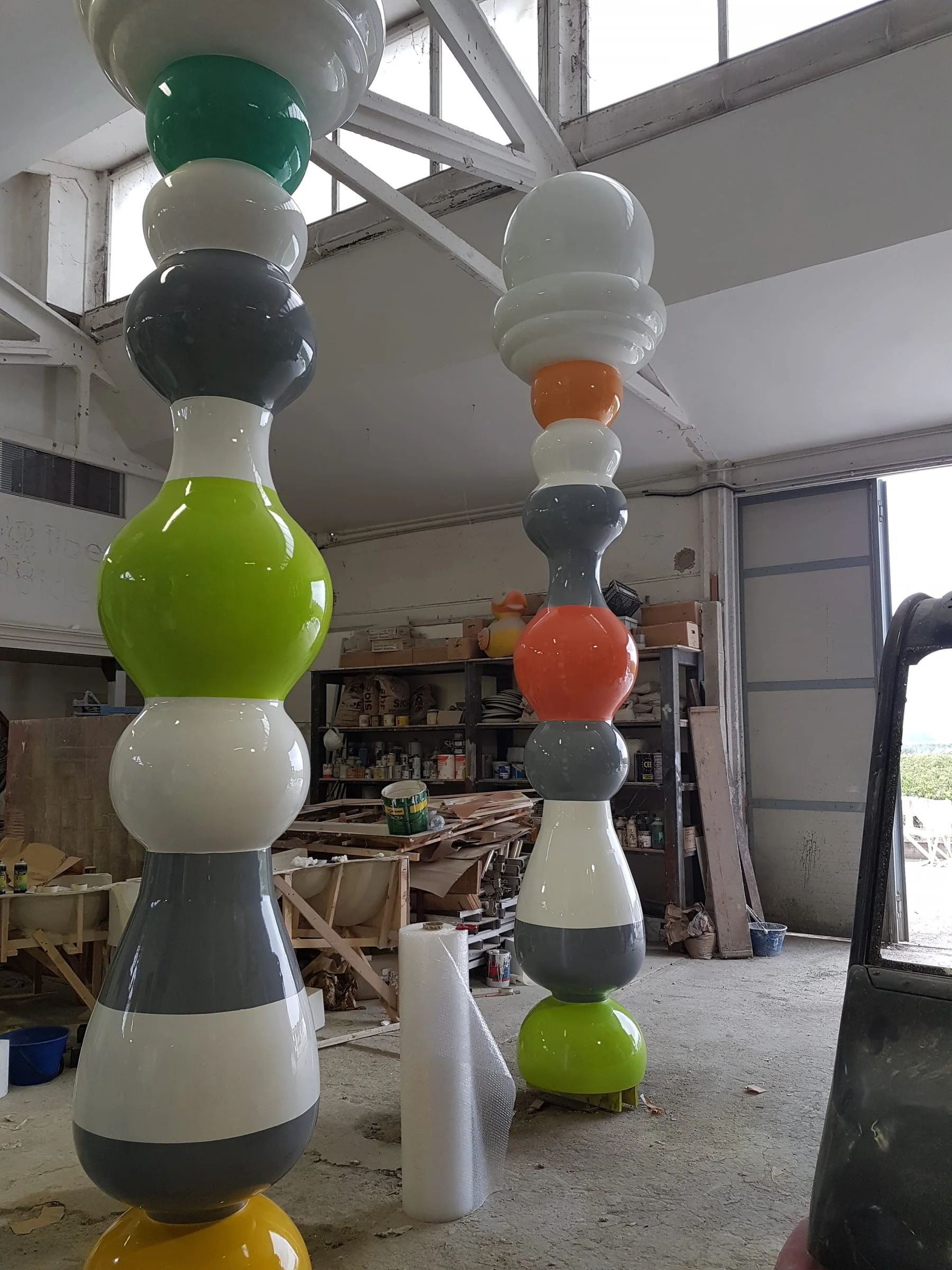 Finished giant pawn-columns in the Creation Art workshops: skittle/balloon-shaped pieces about four metres tall, painted in polychrome stripes — green, orange, grey, yellow, white — arranged horizontally for the final patina, industrial workshop in the background with materials and tools