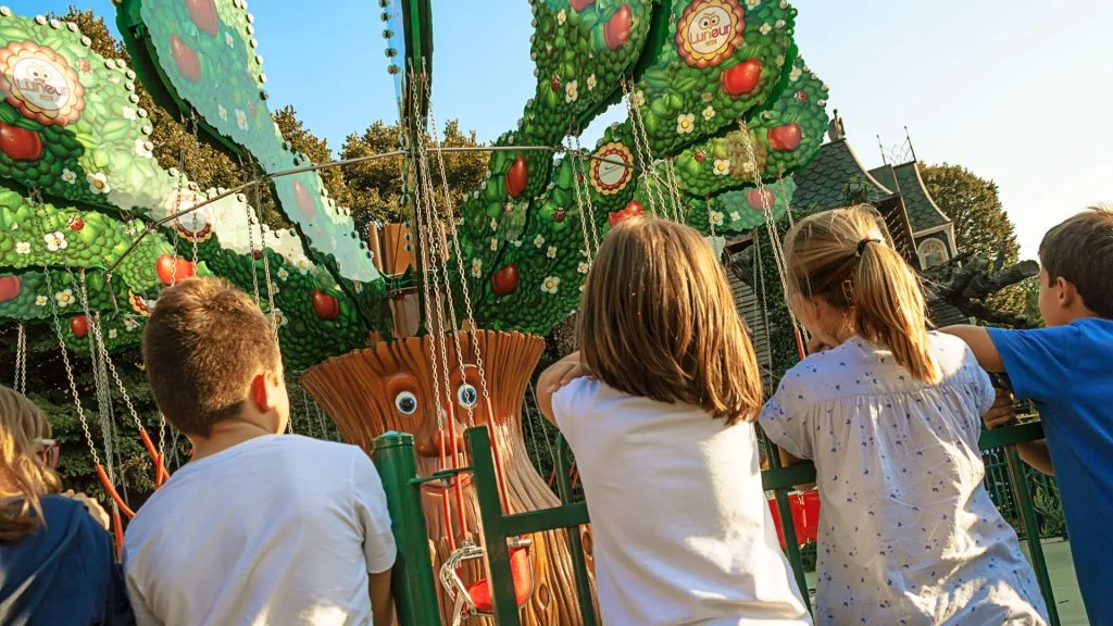 Children seen from behind watching the 'Il Grande Alberone' attraction at Luneur Park: anthropomorphic tree with face, green leaves and red apples, chains of the ride coming down from the branches