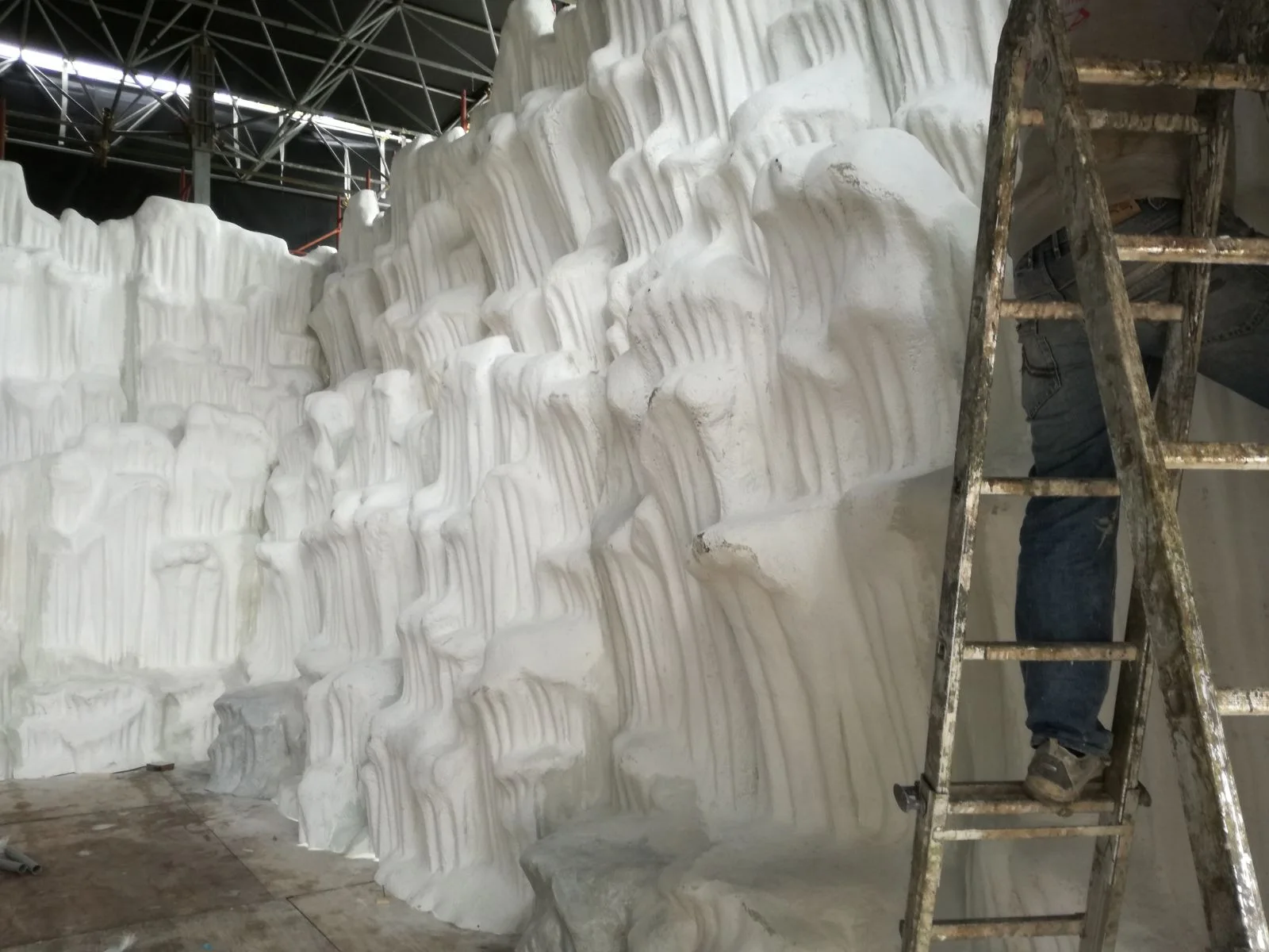 Cantiere della miniera di ghiaccio nel laboratorio Creation Art per il Villaggio di Natale Luneur: grande parete in polistirolo bianco grezzo modellata a stalattiti e formazioni rocciose, scala metallica appoggiata sul lato destro con un operaio sopra in tuta blu, impalcatura visibile sopra