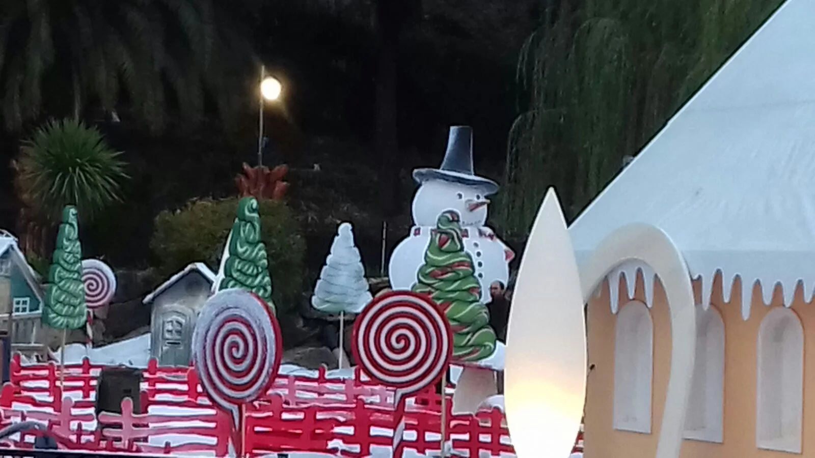 Villaggio di Natale del Luneur installato il 16 novembre 2017: pupazzo di neve alto due metri e mezzo con cilindro grigio, sciarpa rossa e naso a carota arancione al centro, tre alberi di Natale verdi cartoon a spirale ai lati, fila di lecca-lecca a spirale rosso e bianco in primo piano, casetta gingerbread in giallo crema sulla destra, palme illuminate e cielo blu del primo crepuscolo