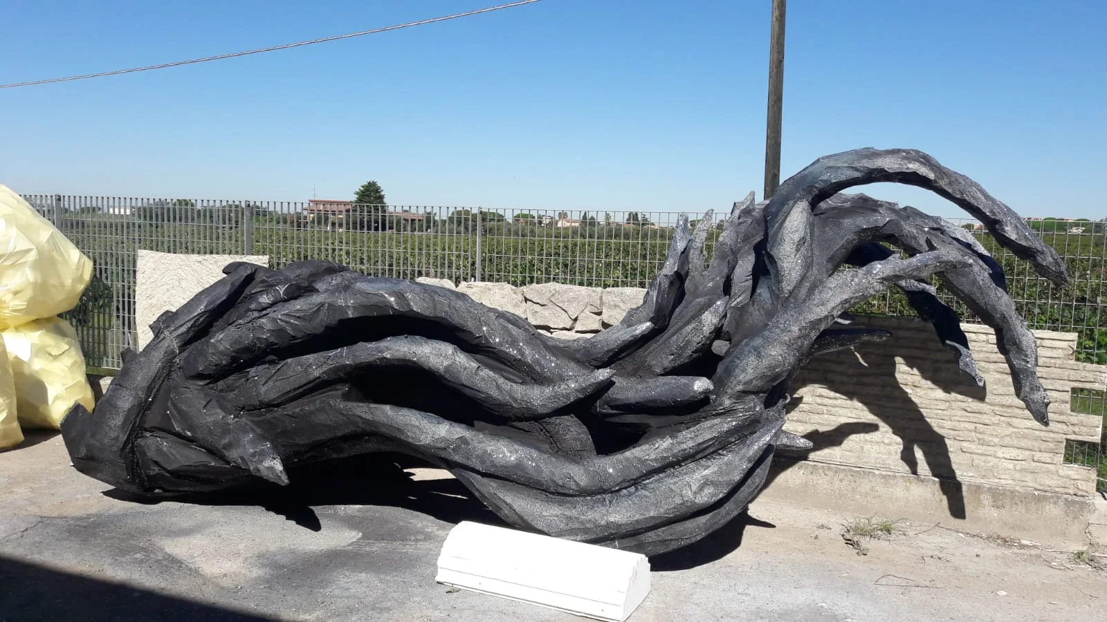 Close-up detail of the black sculpted roots at the workshop: a sculpture of gnarled, intertwined branches like a surge of brambles, black-anthracite finish, laid horizontally on the yard, behind it the metal fence and the fields of the Lazio countryside