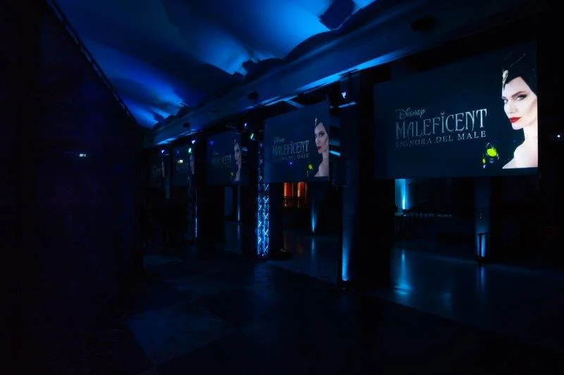 The foyer corridor lined with promotional posters for the film: a series of MALEFICENT MISTRESS OF EVIL panels with a close-up of Angelina Jolie wearing the horn, backlit with cold blue light, dark floor, dark fairytale atmosphere