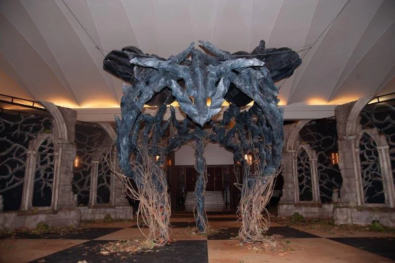 The dragon seen frontally: symmetrical profile of sculpted branch-bones, two Gothic tombstones with pointed arches on either side framing the arcade, black-and-white chequered floor, autumn leaves scattered, white foyer ceiling