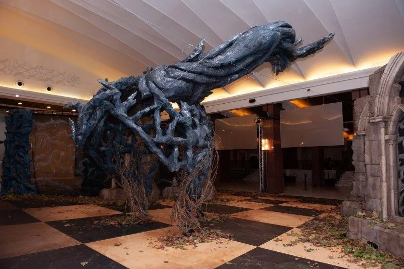 The Maleficent dragon installed at the foyer entrance: blue-grey sculpture about eight metres long, wings spread suspended above the black-and-white chequered floor, two Gothic tombstones in faux stone on either side like a medieval cemetery grave, autumn leaves scattered on the floor, white foyer ceiling lights above