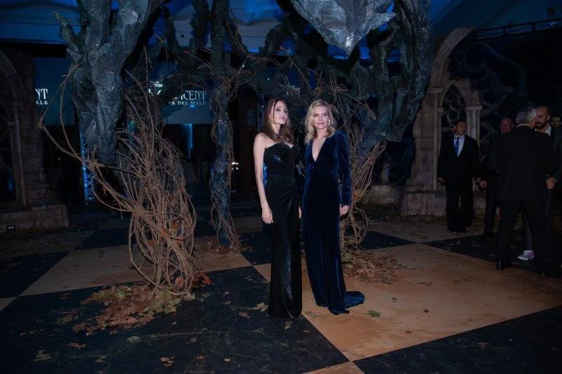 Angelina Jolie and Michelle Pfeiffer posing on the red carpet of the Maleficent European premiere: Jolie in a long black dress on the left, Pfeiffer in midnight-blue velvet on the right, behind them the arch of black sculpted roots and the dragon at the foyer entrance, black-and-white chequered floor, autumn leaves scattered