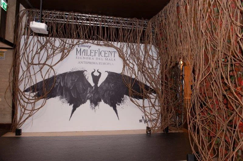 The red carpet photowall: white wall with a central black silhouette of Maleficent (pointed horn on her head and wings spread like a bird), gold lettering 'DISNEY MALEFICENT MISTRESS OF EVIL — EUROPEAN PREMIERE' at the top, framed by a vault of dry branches interwoven by hand above the wall