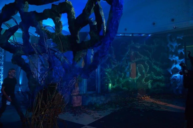 The interior of the European premiere foyer bathed in cold blue light: on the left a large black root-bramble sculpture spiralling from floor to ceiling, behind it green projections of brambles moving across the pale walls, an attendant on the left walking through the space