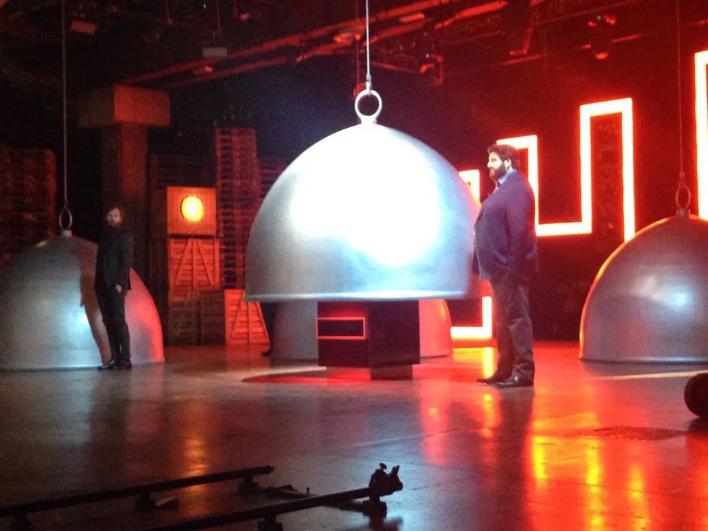 Masterchef Italia set with three large giant suspended silver cloches, lit by warm stage light, beneath the cloches cube-bases with a red luminous band; on the left the judge Bruno Barbieri in a dark jacket, on the right Joe Bastianich in a blue suit; in the background stacked wooden pallets and a red neon luminous Greek-key sign