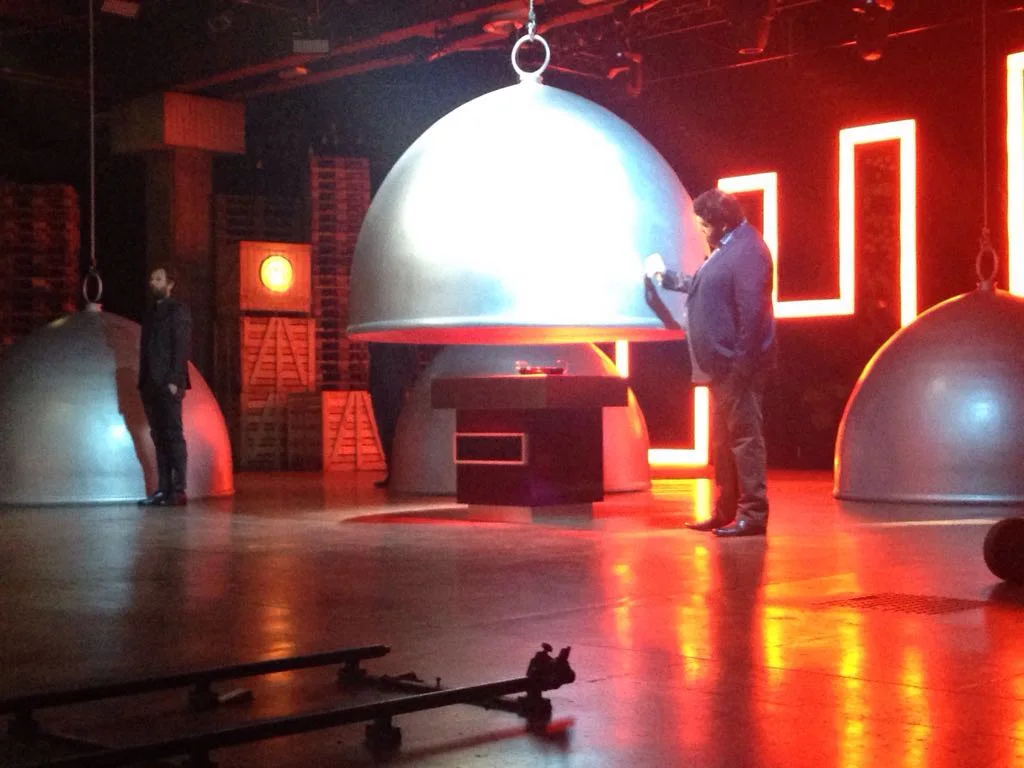 Close-up view of the set with the silver cloche open to reveal the dish below: judge Joe Bastianich's hand lifts the metal cover, detail of the cube-base with red luminous band, behind it the red neon luminous Greek-key sign
