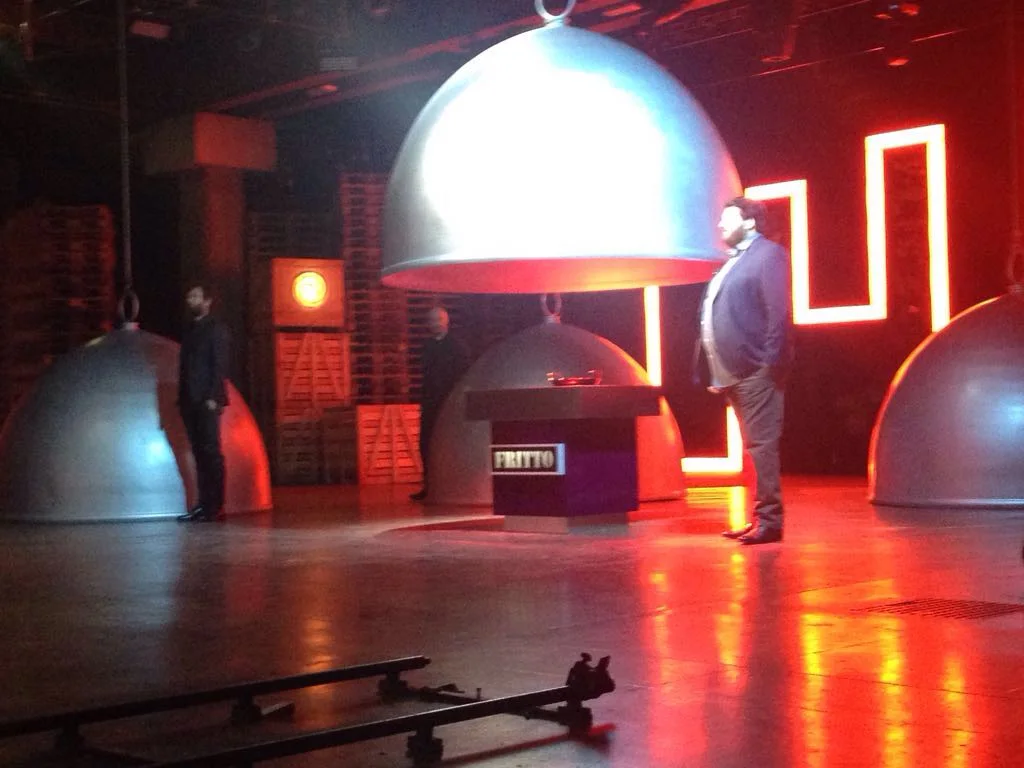 Masterchef set in full action with three contestants half-hidden by the cloches; the central cube shows 'FRITTO' on the red luminous band; on the right Joe Bastianich watches, tense atmosphere with dramatic red background lighting