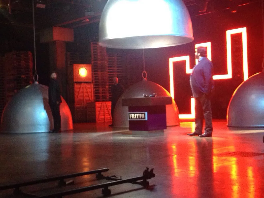 Front view of the set with the central cube reading 'FRITTO' on a red luminous band, beneath the large suspended silver cloche; on either side two more cloches; in the background stacked wooden pallets and the Greek-key red neon luminous sign, TV studio atmosphere
