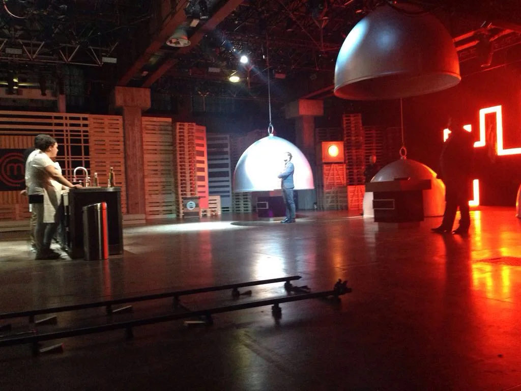 Wide view of the set with a contestant at the vintage kitchen counter on the left, stylised MasterChef logo with red M on the back wall; in the centre the large suspended silver cloche with a contestant standing beneath; stacked wooden pallets as backdrop, professional TV studio lighting