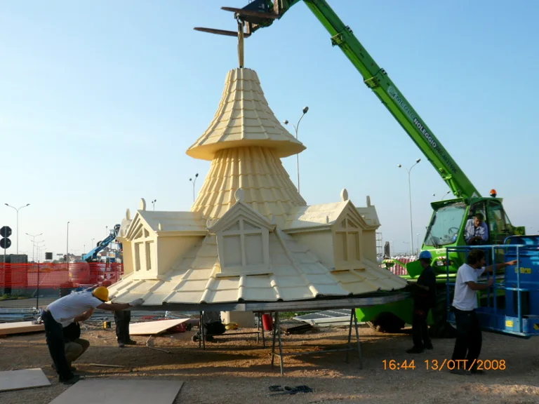 A white onion-shaped tiled dome with spire, lifted by a green 'Holegno' telescopic crane in the construction yard. Workers on the ground guide the movement. Apulian sunset, raised dust