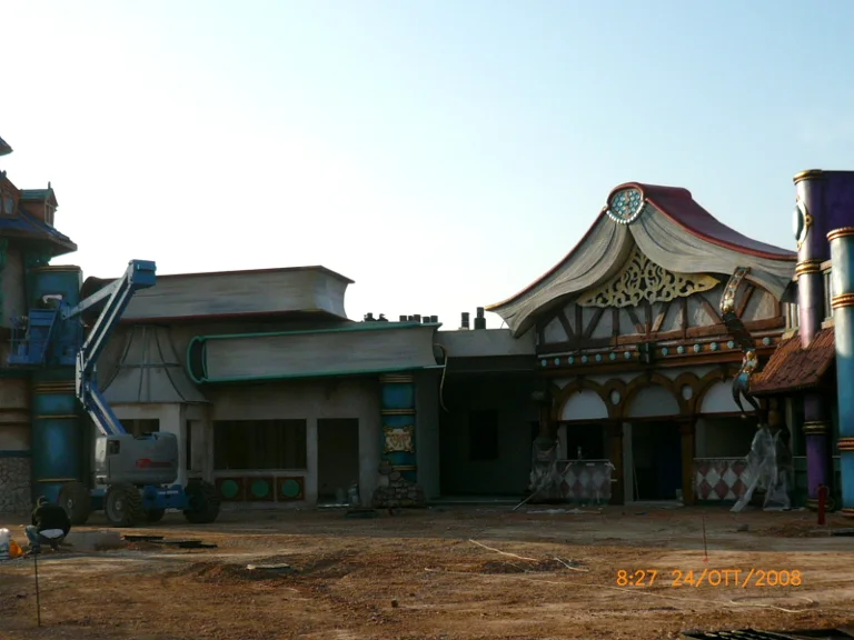 Wide-angle view of the park site during cladding: two themed buildings under construction, scaffolding with a cage on the left, ground still sandy, workers at the back of the scene
