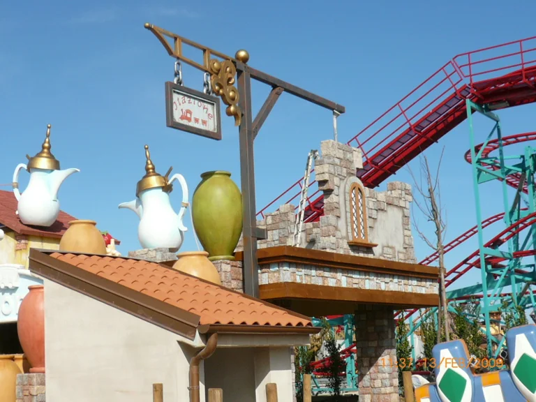 Roof of a shack with two giant white coffee pots on top, one green, a large green terracotta amphora. In the background the red-green structure of the roller coaster.