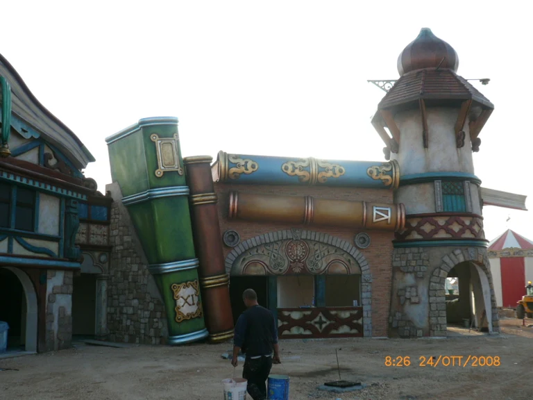 Park facade with two large giant horizontal cannons — one green and one blue — suspended above a red building sculpted as fake stone, gilded decorations, tower with onion dome on the right. A worker in the foreground walks along the structure