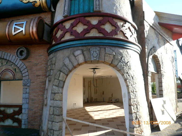 Mid-distance view of a park area with painted cartoon facades, tower buildings with oriental onion domes, bridges, arches. In the background on the right, the red beams of the main roller coaster towering above the themed area