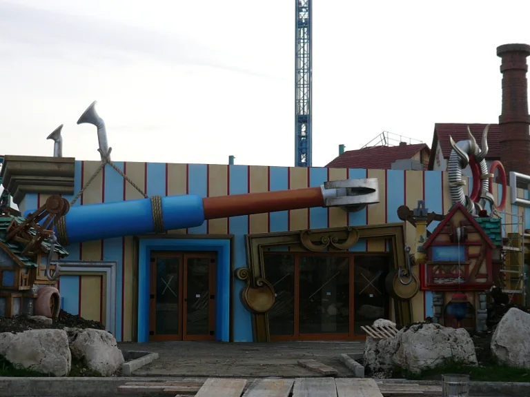 Building with two giant shoes on the roof and a giant horizontal cannon as a lintel. Mustard-colored striped facade, double wooden door. Likely pirate area.