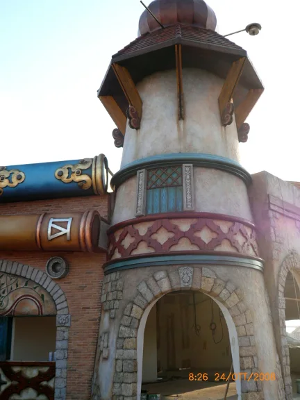 Finished octagonal Mediterranean-style tower with copper onion dome and tiled roof. At its base a large decorated horizontal blue cannon. Cream-coloured walls sculpted to look like stone