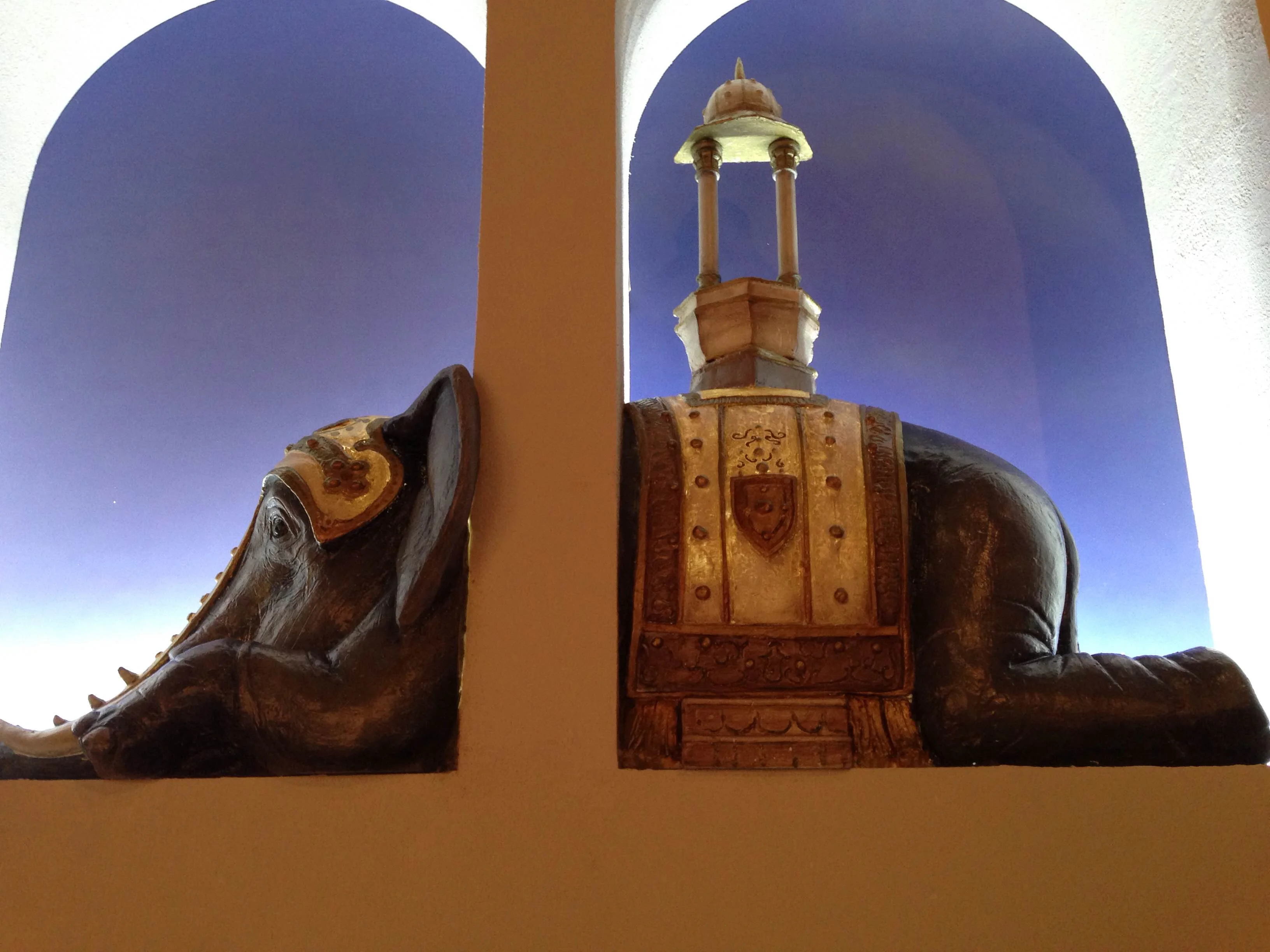 The same elephant seen through two large arches of the installation: the muzzle in profile on the left in a niche, the back with ceremonial pavilion on the right in another arch, dark blue-violet background of the nocturnal cyclorama