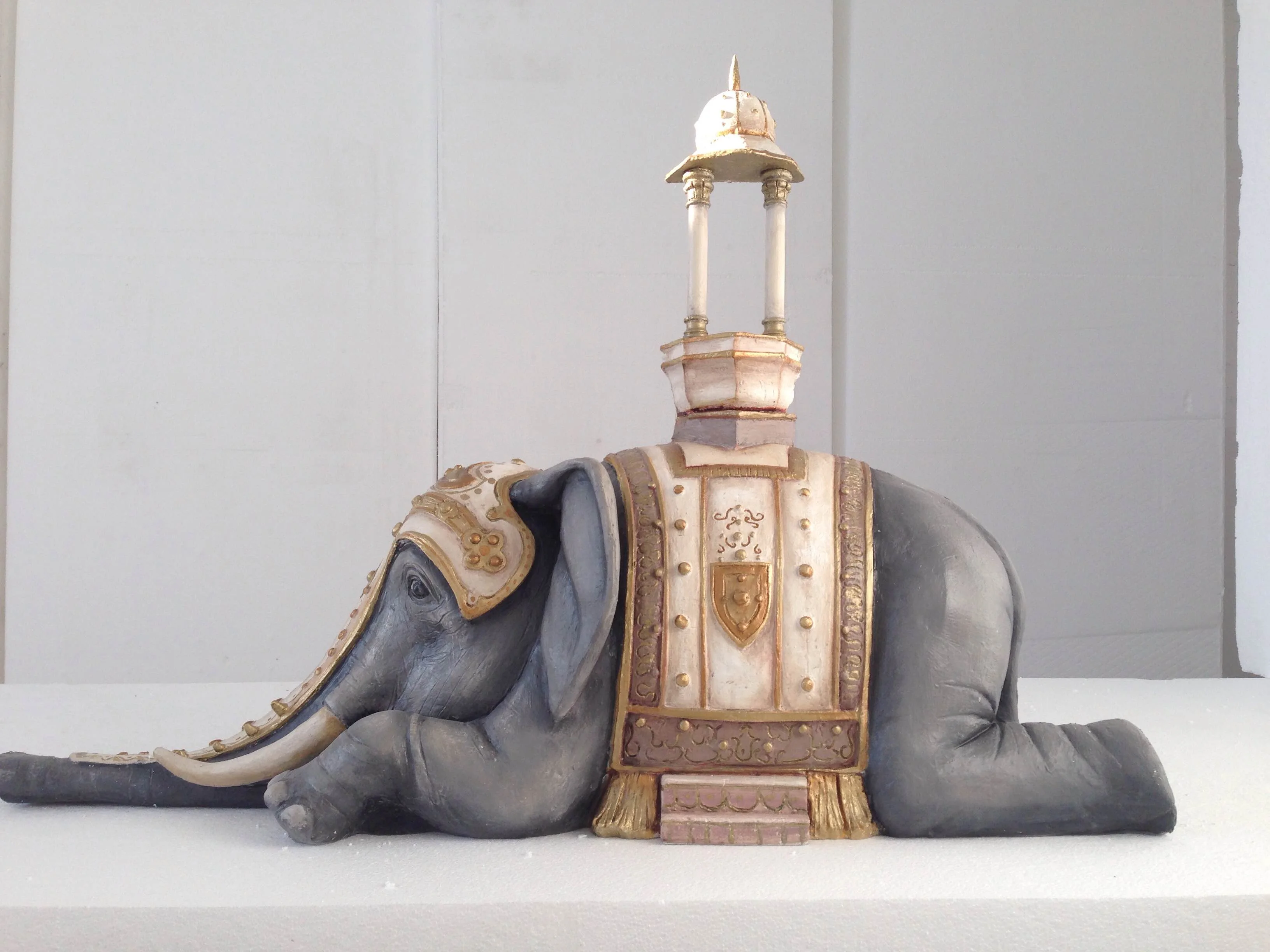 Elephant sculpted in the round for the Fellini Exhibition: animal's body resting on the ground in grey clay, ceremonial harness in faux gilded sheet metal on the flanks with central crest, small pavilion/shrine on its back with pointed dome and flag, full side view