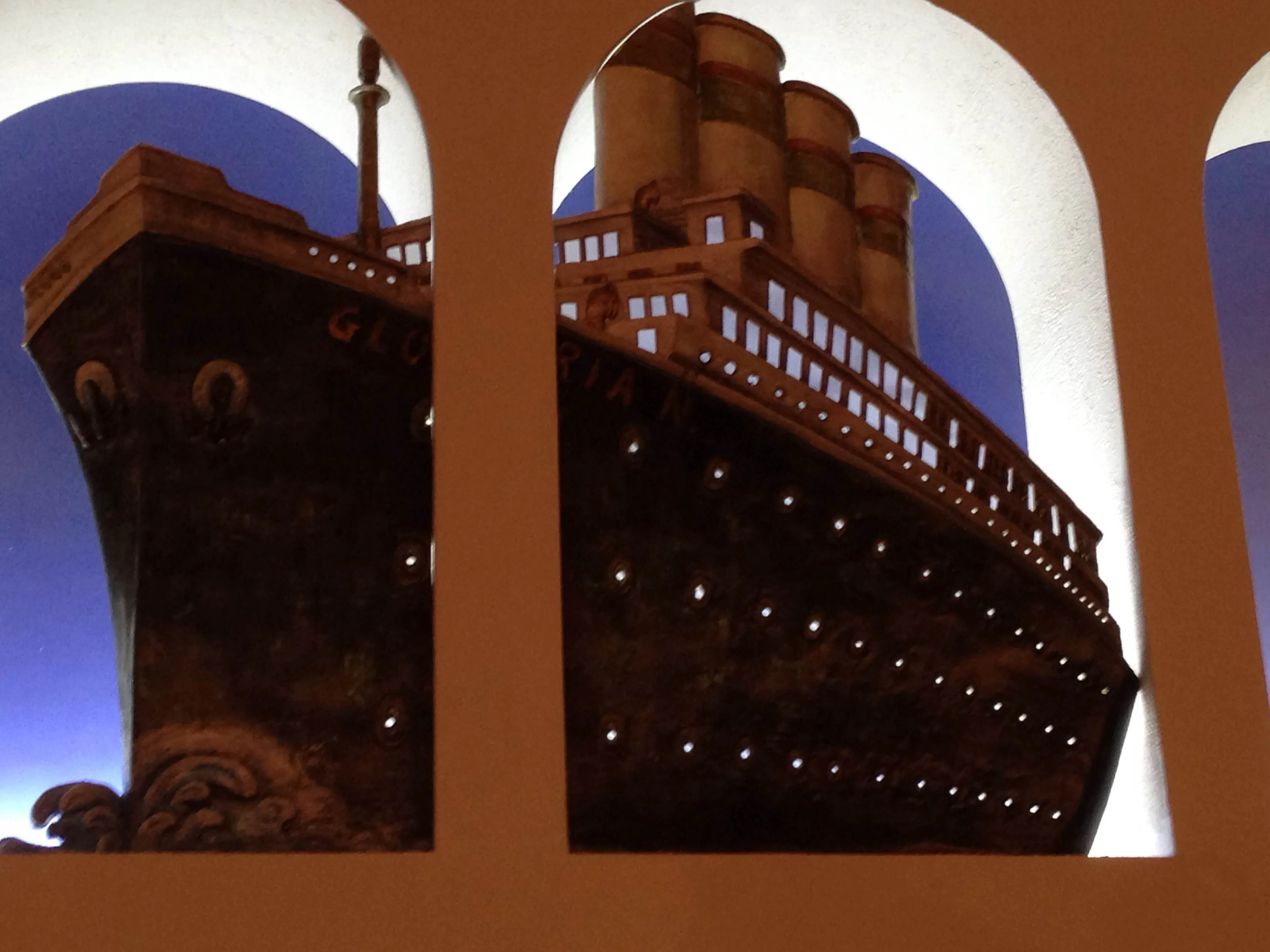 The ocean liner Rex from Amarcord rebuilt as scenography for the Fellini Exhibition at Macro La Pelanda: dark bow of the ship with the inscription 'GLORIA' on the side, three large red funnels, row of illuminated white portholes along the hull, midnight blue background; the scene is seen through two large beige arches of the museum that frame the ship like a painting